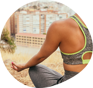 Person in athletic clothing sits cross-legged outdoors, meditating with one hand on knee. City buildings are visible in the blurred background.