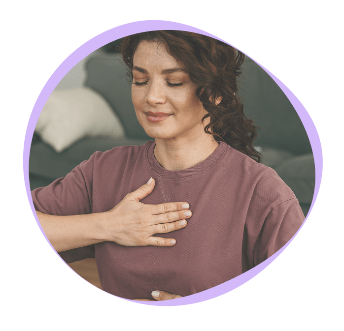 Woman with dark hair sits indoors, eyes closed, and places one hand on her chest, appearing calm and relaxed.