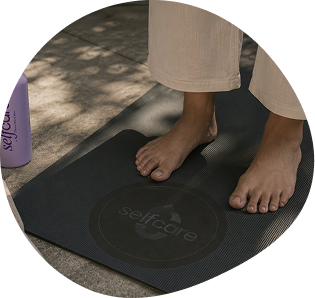 Bare feet stand on a black yoga mat with “selfcare” written on it; a purple water bottle with “selfcare” branding is nearby.