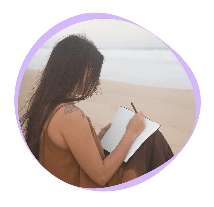 A woman with long hair sits on a beach, writing in a notebook with a pen. The ocean is visible in the background.