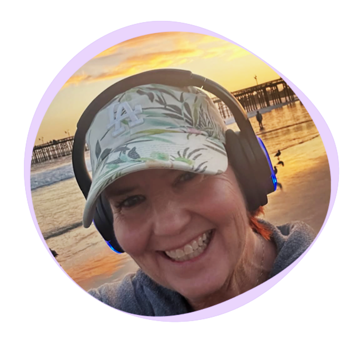 Woman wearing a floral cap and headphones smiles at the beach during sunset, with a pier and seagulls in the background.