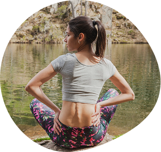 Woman in athletic wear sitting by a lake, facing away, with hands on her lower back as if experiencing discomfort.