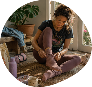 Woman sitting on a woven rug indoors, putting on socks, surrounded by a yoga mat, water bottle, mug, and potted plant. Sunlight streams in through glass doors behind her.